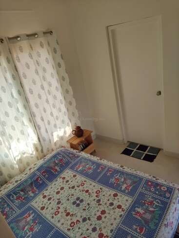 This cozy bedroom features a colorful, patterned bedsheet, white printed curtains, a small wooden side table with books and a teapot, and a simple white door.