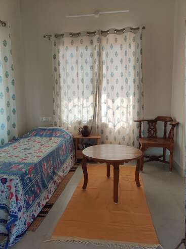 A cozy room with a single bed, colorful patterned bedspread, wooden round table, chair, orange rug, sheer curtains, side table, and a decorative clay pot.