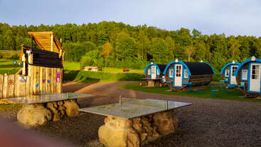 Das Bild zeigt einen sonnigen Campingplatz mit einzigartigen tonnenförmigen Hütten, Picknicktischen, steinernen Tischtennisplatten, einem hölzernen Unterstand, grünem Gras und einem üppigen Wald im Hintergrund.