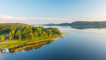 Eine ruhige Seeszene mit üppig grünen Bäumen, die das Ufer säumen und sich im ruhigen Wasser spiegeln. In der Ferne erheben sich Hügel unter einem klaren blauen Himmel. Friedliche Atmosphäre.