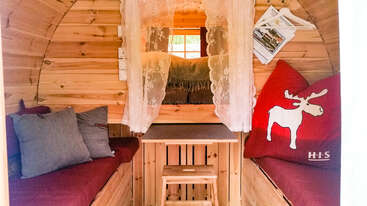 In dieser gemütlichen Holzhütte gibt es zwei rote gepolsterte Bänke, einen kleinen Tisch, Spitzenvorhänge, weiche Kissen und ein Fenster, durch das natürliches Licht einfällt.