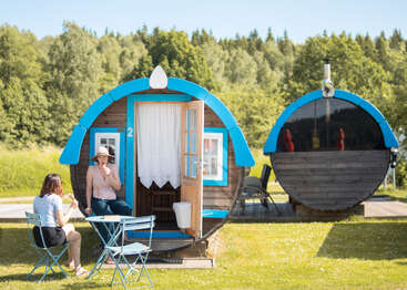 Zwei Frauen sitzen an einem kleinen Tisch vor einer bezaubernden, tonnenförmigen Hütte mit blauer Verkleidung und genießen einen sonnigen Tag inmitten von üppigen grünen Bäumen und Gras.