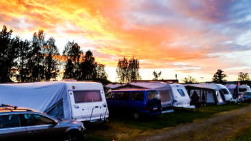Unter einem atemberaubenden, farbenfrohen Sonnenuntergangshimmel, umgeben von Bäumen, sind Reihen von Wohnwagen und Fahrzeugen auf dem Gras geparkt und schaffen eine friedliche Campingszene mit lebendigem Licht.