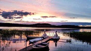 Ein ruhiger See bei Sonnenuntergang mit schwanenförmigen Tretbooten, einem hölzernen Steg und einem Ruderboot, umgeben von Schilf unter einem bunten, dramatischen Himmel. Friedliche Szene.