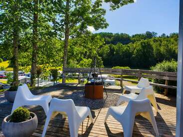 Dieses Bild zeigt eine sonnige Holzterrasse mit modernen weißen Stühlen, die um eine Feuerstelle herum angeordnet sind, umgeben von Bäumen und mit Blick auf eine grüne, malerische Landschaft.