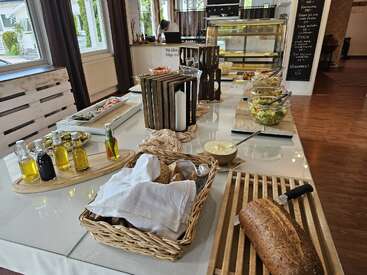 Auf einem Buffettisch stehen frisches Brot, Olivenöle, Salate, Aufstriche und verschiedene kalte Speisen bereit – in einem hellen, gemütlichen Speisesaal mit rustikaler Einrichtung und Holzakzenten.