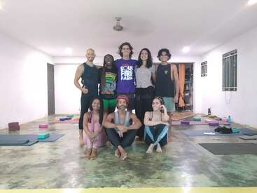 Sept personnes posent ensemble dans un studio de yoga, certaines debout, d'autres assises les jambes croisées. Des tapis de yoga, des blocs et un environnement serein et minimaliste suggèrent un cours collectif.