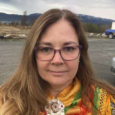 Das Bild zeigt eine Frau mit langen braunen Haaren und Brille, die einen leuchtend gelben Schal mit einer großen Brosche trägt, vor einer malerischen Kulisse aus Bergen und einem bewölkten Himmel.