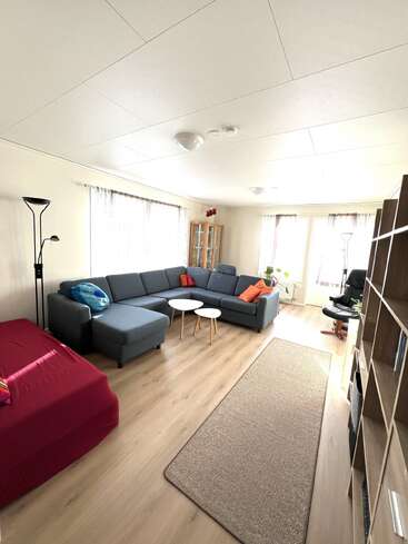 Dieses helle Wohnzimmer verfügt über ein großes graues Sektionssofa, bunte Kissen, zwei runde Tische, ein rot bezogenes Bett, Holzregale und viel Tageslicht.