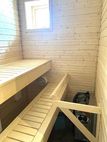 Dieses Bild zeigt einen kleinen hölzernen Saunaraum mit hellen Wänden, zwei Bankebenen, einem Fenster, einem Ofen mit Steinen und einer schützenden Holzbarriere.