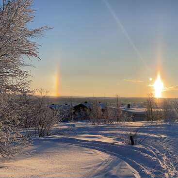 Eine verschneite Landschaft unter klarem Himmel, mit frostigen Bäumen, Häusern und einem leuchtenden Sonnenhund-Phänomen mit strahlenden, bunten Lichtsäulen bei Sonnenaufgang oder Sonnenuntergang.