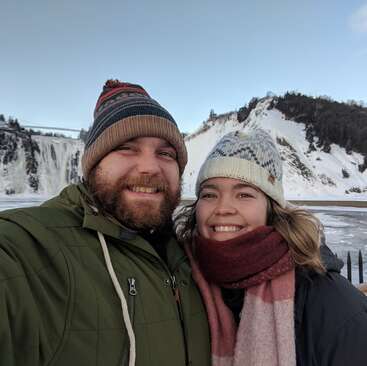 L'image représente un homme et une femme posant pour un selfie devant une montagne enneigée et une cascade, tous deux portant des bonnets et des manteaux d'hiver.