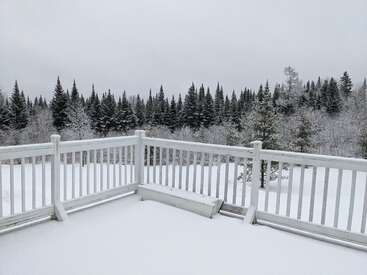 L'image représente une scène hivernale sereine depuis une terrasse, avec une balustrade couverte de neige et un paysage enneigé avec des arbres en arrière-plan sous un ciel couvert.