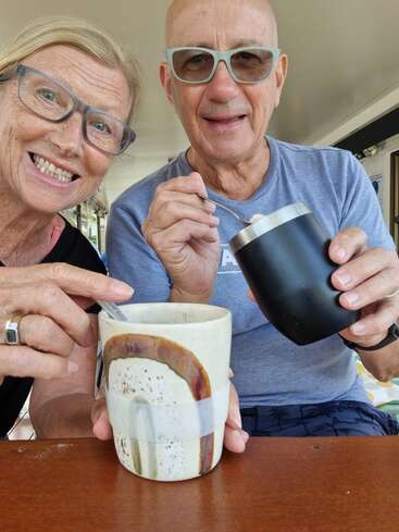 Um casal de idosos sorridente, de óculos, está sentado lado a lado, segurando xícaras de café e colheres, aproveitando um momento de descontração em uma mesa num ambiente claro e alegre.