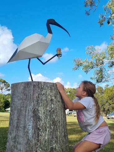 Uma menina de bermuda rosa olha para cima e sorri para uma grande escultura de pássaro de metal segurando uma casquinha de sorvete, empoleirada no topo de um toco de árvore ao ar livre.