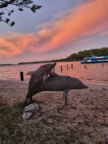 Duas esculturas de golfinhos de metal numa praia de areia ao pôr do sol, com um céu colorido, árvores ao longe, um barco azul atracado e águas calmas ao fundo.
