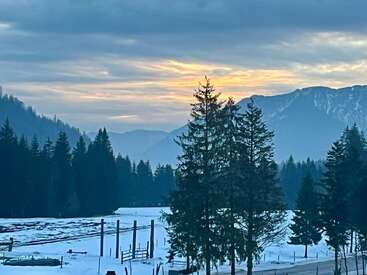 Das Bild zeigt eine verschneite Landschaft bei Sonnenuntergang mit hohen immergrünen Bäumen, fernen Bergen und einem dramatischen, wolkenverhangenen Himmel, der in sanftem orangefarbenem Licht erstrahlt. Friedliche Szene.