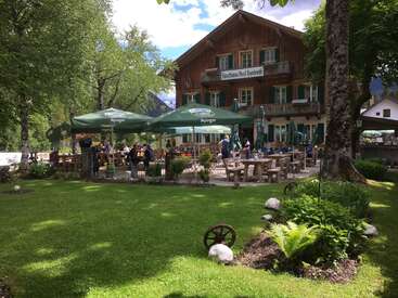 Ein charmantes, rustikales Gästehaus mit Sitzplätzen im Freien unter grünen Sonnenschirmen, umgeben von üppigem Grün, Bäumen und einem gepflegten Garten. Die Menschen genießen einen ruhigen Nachmittag.