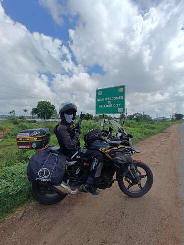 A motorcyclist, fully geared, poses next to a touring bike on a rural road. A green sign reads "NHAI Welcomes You to Nellore City."