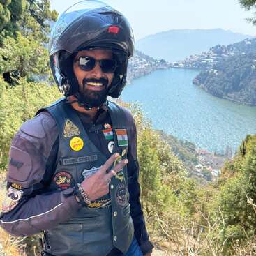 A biker in a helmet and sunglasses smiles while flashing a peace sign, standing before a scenic lake surrounded by green hills and distant mountains.