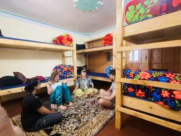 Four people sit on a patterned carpet in a cozy dorm room with wooden bunk beds and colorful floral blankets, chatting and enjoying each others' company.