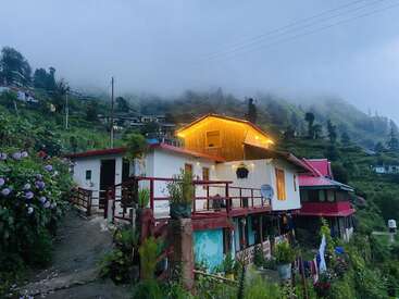 Una encantadora casa en la ladera de una colina con luces resplandecientes se asienta en medio de exuberante vegetación y montañas neblinosas, rodeada de flores y naturaleza vibrante, creando un ambiente tranquilo y acogedor.