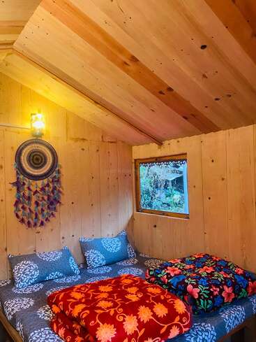 Une chambre confortable dans une cabane en bois avec un plafond incliné, un attrape-rêves sur le mur, une literie à motifs, des couvertures colorées, une fenêtre et une lampe à la lueur chaleureuse.
