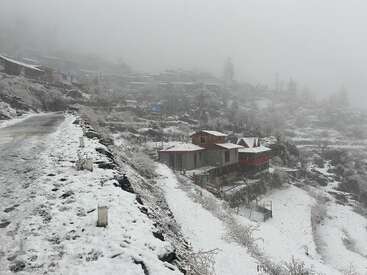 Une scène hivernale brumeuse montre un village enneigé niché à flanc de colline, avec des petites maisons éparpillées, et une route calme et glacée qui serpente à travers le paysage.