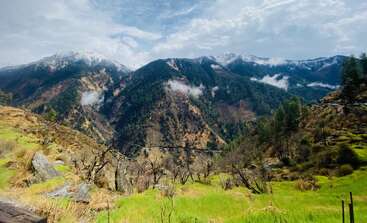 L'image présente un paysage de montagne à couper le souffle, avec des vallées verdoyantes, des arbres épars et des sommets enneigés. Des vagues de nuages s'accrochent joliment entre les pentes boisées.