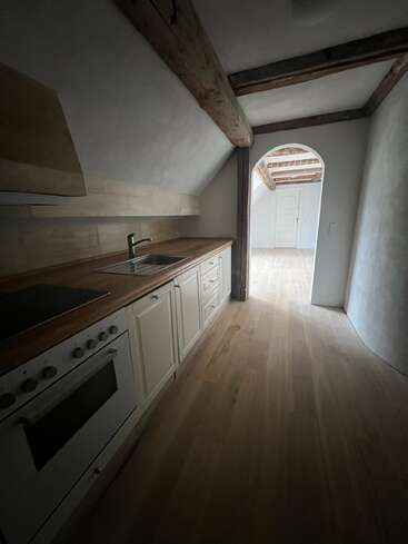 Cette image montre une cuisine étroite avec un sol en bois, des armoires blanches, des poutres apparentes, un four et un évier, menant à une porte voûtée lumineuse.