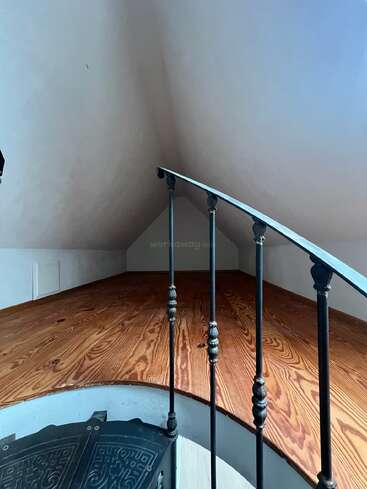 Cette image montre un petit grenier ou loft avec des planchers en bois, des murs blancs inclinés et une rampe en fer noir, vu du haut d'un escalier.