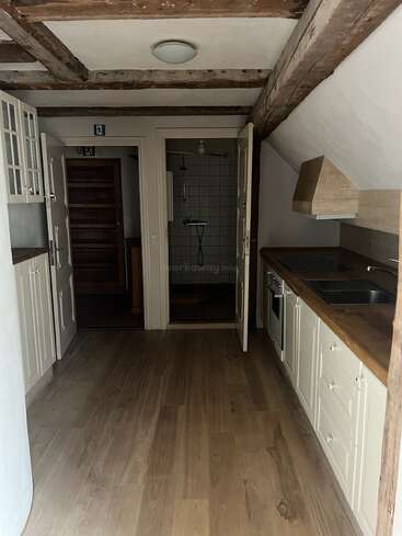 Cette image montre une cuisine étroite avec des sols en bois, des armoires blanches, des poutres apparentes au plafond et des portes menant à une salle de bains aux murs carrelés.