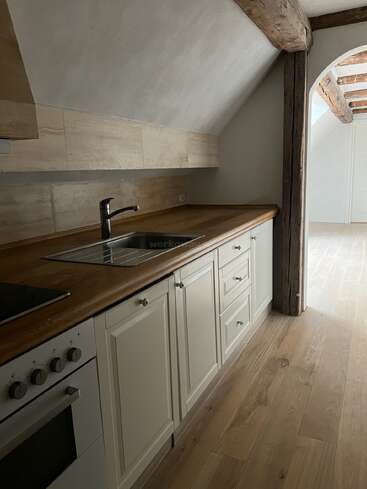 Cette cuisine rustique présente des poutres en bois, un plafond incliné, des appareils modernes, des armoires de couleur crème, des plans de travail en bois, un évier en acier inoxydable et une lumière naturelle qui renforce l'atmosphère chaleureuse et accueillante.