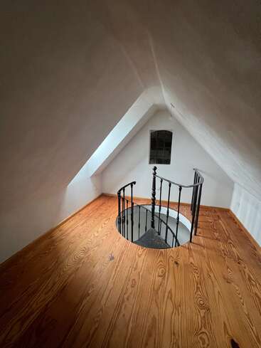 Cette image montre une pièce mansardée avec des murs blancs inclinés, un plancher en bois, un escalier en colimaçon, une petite fenêtre et une lucarne qui apporte de la lumière naturelle. Un espace confortable.