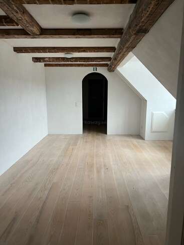 Cette image montre une pièce vide et lumineuse avec des planchers en bois, des murs blancs, des poutres apparentes au plafond et une porte voûtée menant à un espace plus sombre.