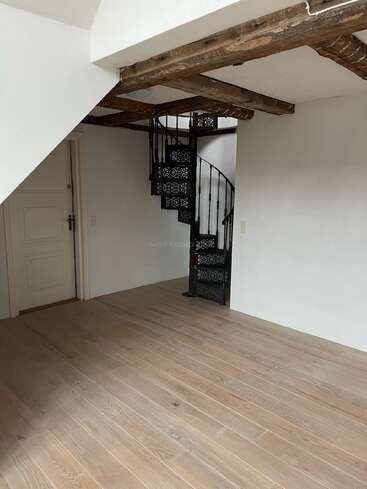 Cette image montre une pièce vide avec un sol en bois clair, des poutres apparentes au plafond, des murs blancs et un escalier en colimaçon noir décoratif près d'une porte fermée.