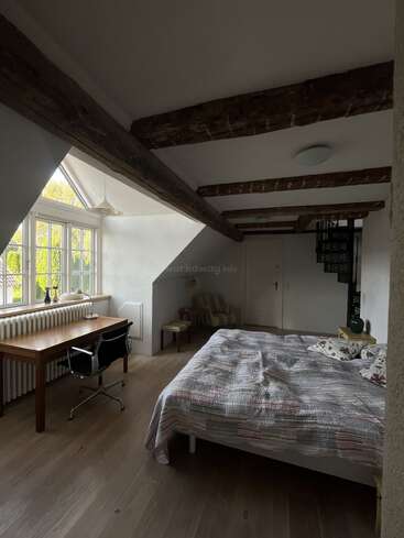 Cette chambre mansardée confortable comprend des poutres en bois, un lit double avec des draps à motifs, un bureau en bois près d'une grande fenêtre, une chaise et un escalier en colimaçon.
