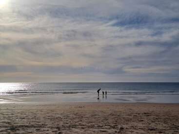 A serene beach scene at sunset with calm waves, cloudy sky, and three figures standing by the water, reflecting on the peaceful, open shoreline.