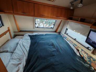 This cozy camper van bedroom features a double bed with white pillows, blue blanket, surrounding windows, upper storage cabinets, shelving, and a small wall-mounted television.