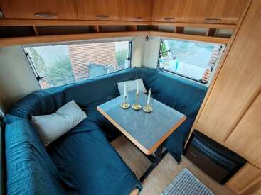 This image shows a cozy caravan dining area with a U-shaped blue couch, two pillows, a small table, three candlesticks, large windows, and wooden cabinets.