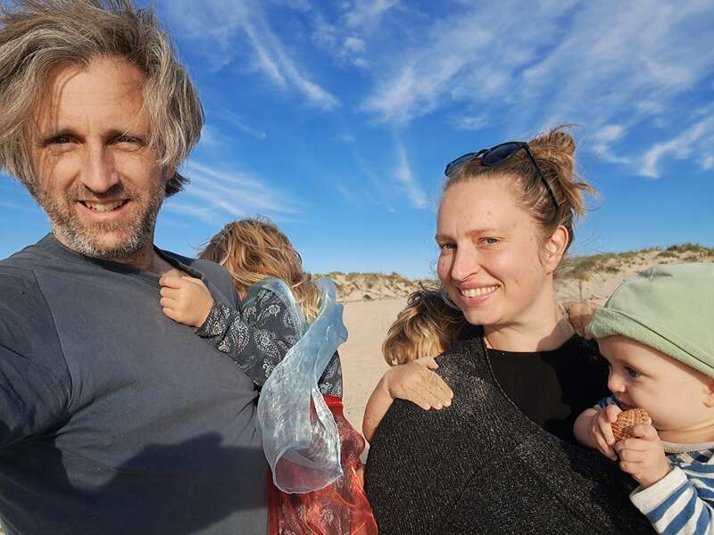 A family of four poses for a selfie at the beach, smiling under a bright blue sky, with sand dunes and wavy clouds in the background.