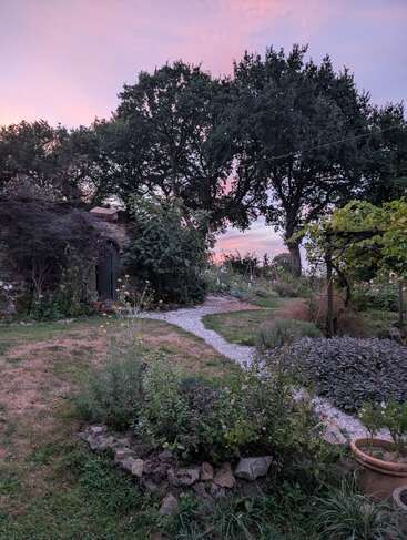 Una tranquila escena de jardín con sinuosos senderos de grava, exuberante vegetación, parterres de flores, árboles altos y un cobertizo rústico bajo un suave cielo vespertino de colores pastel.