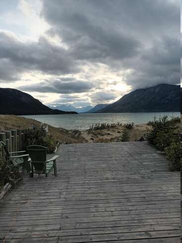 Una terraza de madera con dos sillas verdes da a un sereno lago rodeado de montañas, bajo un cielo nublado y espectacular. Los pinos y las dunas de arena completan la tranquila escena.