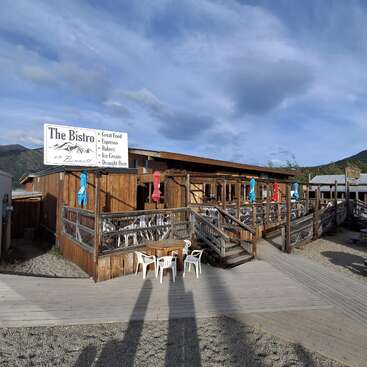 A rustic wooden bistro sits under a blue sky, featuring outdoor seating, colorful umbrellas, and a sign advertising great food, espresso, bakery, ice cream, and beer.