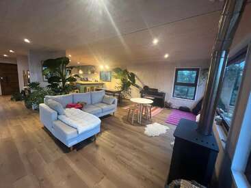 This cozy living room features a light sectional sofa, wood stove, large plants, round table with stools, piano, wooden floors, and an open kitchen area.