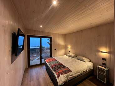 This cozy wooden bedroom features a large bed with colorful bedspread, soft lighting, wall-mounted TV, sliding glass doors, and a scenic view outside. Warm, inviting atmosphere.