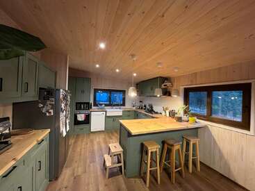 This is a cozy wooden kitchen with green cabinets, an island with stools, modern appliances, pendant lights, large windows, and warm, inviting natural finishes throughout.