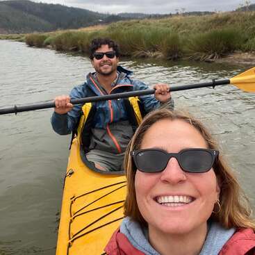 A smiling woman and man are kayaking together on a river, both wearing sunglasses and jackets, surrounded by nature, grassy banks, and cloudy skies.