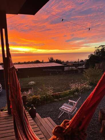 A cozy porch with a hammock overlooks a peaceful garden and distant ocean, illuminated by a magnificent, vibrant orange and pink sunset sky. Tranquil, inviting scene.
