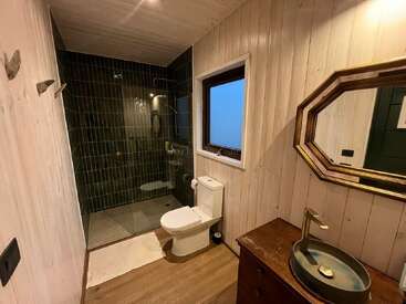 This bathroom features wood-paneled walls, a modern toilet, a glass-enclosed shower with dark tiles, a rustic vanity with a vessel sink, and a framed mirror.